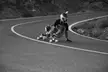 A person with tattoos wearing a helmet and streetwear longboarding down a winding road in black and white.