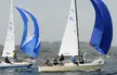 Two J-24 sailboats with white and blue sails racing on a lake with forested hills and a group of sailors.
