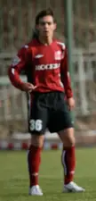Sergei Shudrov, a soccer player with brown hair and fair skin, wearing a red jersey and black shorts on a grass field