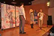 Three people on a wooden stage in front of a bird banner, including a blonde girl in a short grey mini dress.