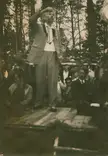 Vydūnas in a suit and tie delivering a funeral speech from a wooden platform to a group in a forest