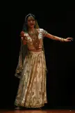 Indian woman in a gold lehenga choli and ornate jewelry performing a dance barefoot on a dark stage