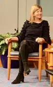 Maya MacGuineas with blonde hair in a black textured dress, tights, and boots sitting in a wooden chair