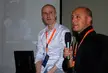 Two bald men with orange lanyards, one in a white shirt and one in a black jacket, at a social media tour in Milano.