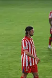 Ujfaluši in a red and white striped Atlético Madrid jersey and headband on a grass soccer field.