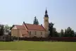 Yellow church with a red roof and tall steeple in Gościce, Nysa County, behind a brick wall and green field