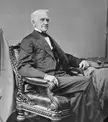 Francis Thomas in a black and white vintage photo, wearing a dark suit and bowtie in an ornate chair