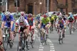 A group of cyclists in colorful athletic wear and helmets racing through a town street with brick buildings.