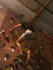 A fair skin woman in a black t-shirt and short shorts climbing an indoor rock wall with a safety harness