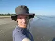 A smiling man with fair skin in a blue t-shirt and grey wide-brimmed hat taking a selfie on a sandy beach by the ocean.