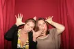 Two fair-skinned women with brown hair making funny faces and gestures in a photo booth with a red curtain
