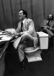 A laughing man in a suit and tie sitting on a toilet in an office setting, a funny work meme.
