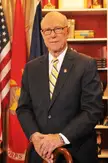 Pat Roberts smiling in a dark pinstripe suit, white shirt, and striped tie in an office with flags.
