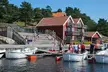 People in swimwear and life vests relaxing by red houses and white boats on the Tregde waterfront.