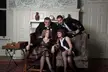 Four friends in a vintage living room with floral print wallpaper, a fireplace, and armchairs.