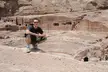 A man in a black t-shirt and sunglasses crouching before the rock-cut amphitheater in Petra.