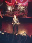 Kanye West in a dark suit and sunglasses performing on a concert stage with a drummer and guitarist.