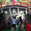 A group of people walking under a red bridge decorated with white lanterns in a scenic Japanese landscape.