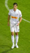 Zoltán Lipták in a white v-neck jersey and shorts standing on a green soccer field