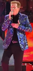 Emmanuel singing into a microphone wearing a blue floral print blazer and black trousers against a red background