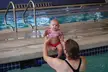 A woman with brown hair in a black swimsuit and a baby in a pink bikini at an indoor swimming pool.