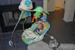 A fair-skinned redhead infant in a blue patterned shirt sitting in a Fisher-Price baby swing near a dog and fireplace