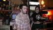 A surprised man in a plaid shirt and a tattooed person in a black t-shirt holding plates of food in a bar.