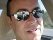 Close-up mirror selfie of a smiling man with dark hair and mirrored sunglasses inside a car interior.