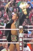 The Miz in a wrestling ring wearing a leather jacket and trunks, holding two championship belts before a crowd.