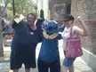 A man and woman in casual clothes smiling next to a person in a blue Stitch costume with a flower lei on a city street.