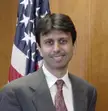 Jindal smiling in a grey suit, white shirt, and red patterned tie in front of a US flag and wooden wall.