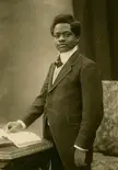 Paul Panda Farnana in a dark suit and white shirt, standing by a table with a book in a vintage black and white photo.