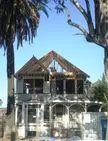 A house under construction with exposed wooden roof framing, palm trees, and a chain-link fence in South Los Angeles.