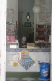 El hombre de las chuches standing behind a counter in a small shop with food menus and a blue bin.