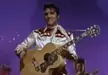Elvis Presley in a white western shirt playing an acoustic guitar against a purple background.