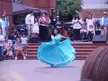 A woman in a large blue off-the-shoulder dress dancing on a brick stage before spectators and musicians.