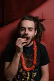 A man with long brown hair in a bun, wearing a black t-shirt and orange lei, eating against a red background.