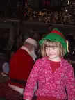 A blonde child in a green elf hat and pink floral cardigan standing with a person in a Santa costume