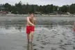 A shirtless man with pale skin in red short shorts standing barefoot on a muddy beach.