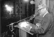 Willy Brandt wearing glasses and a suit, speaking into a microphone at a podium outdoors at night