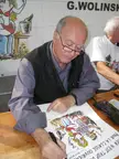 Wolinski wearing glasses and a black vest drawing a cartoon at a wooden table