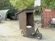 Seniortransportation: a wooden outhouse mounted on a motorized three-wheeled scooter in a gravel area