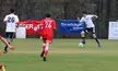 Three soccer players in athletic wear, including one in a red Texans 26 jersey, on a grass field with banners.