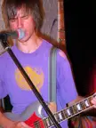 A slender man with brown hair in a purple tie-dye t-shirt singing into a microphone while playing an electric guitar.