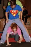 College girls in a group, with a woman in a blue shirt standing over a redhead in a pink shirt on a red mat.