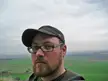 Daniel Sieradski wearing a baseball cap, glasses, and a hoodie in a rural field
