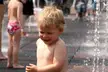 A smiling blonde child in swimwear playing in a water fountain with a group of people.