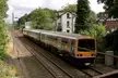 Silver and yellow train passing the original Cheadle Hulme station house and green trees.