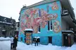 Couple in winter gear standing before a colorful abstract mural on a light blue building in Prince Albert