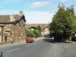 Red car driving past a stone building toward the Cullingworth Viaduct under a blue sky.